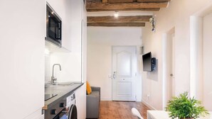 Cocina compacta con microondas moderno, pileta y horno integrado. Un sofá gris está ubicado cerca de una puerta blanca, y una planta en maceta sobre una mesita.