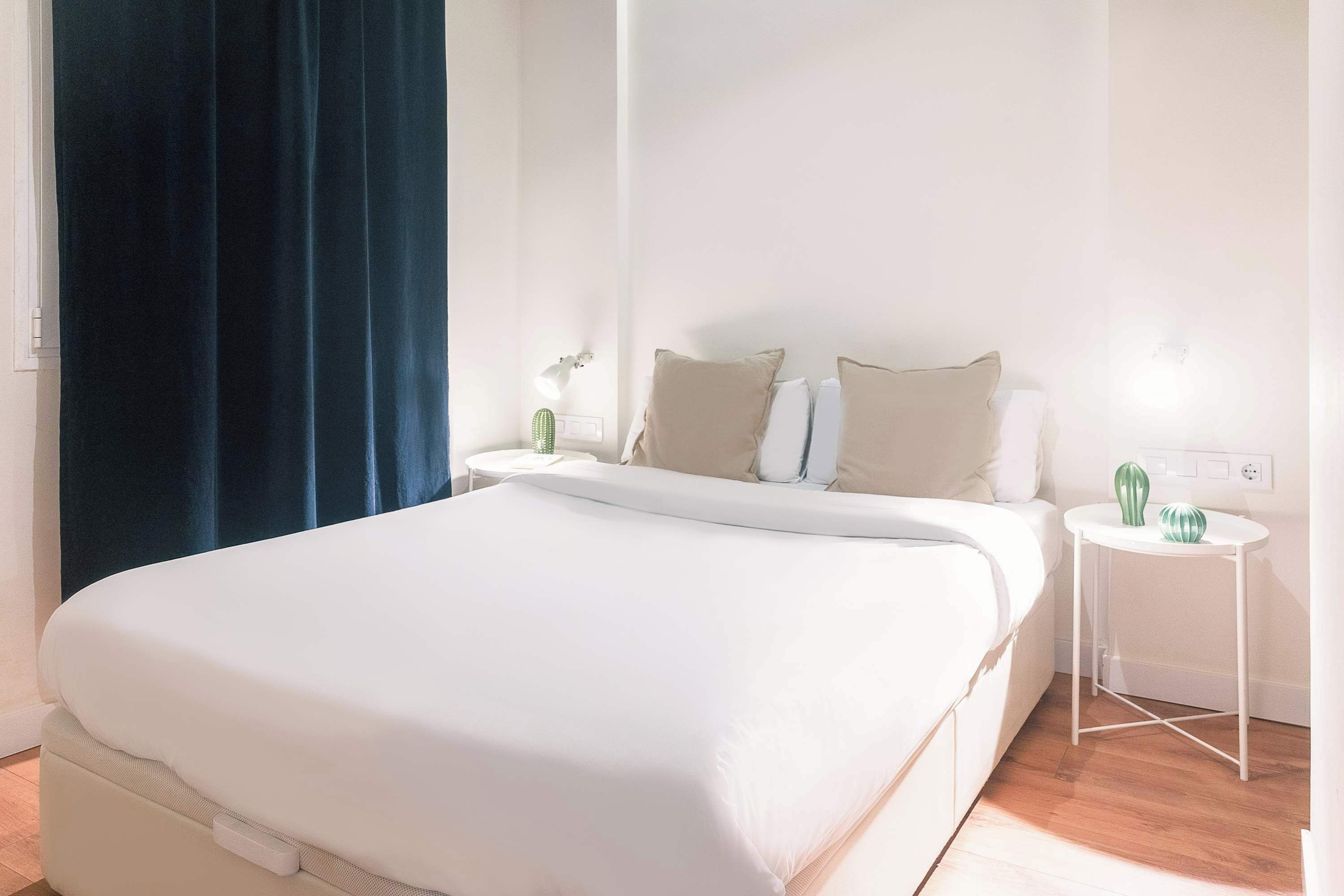 A neatly made bed with a white mattress and pillows, a small white bedside table with a lamp, and a cactus on the table.