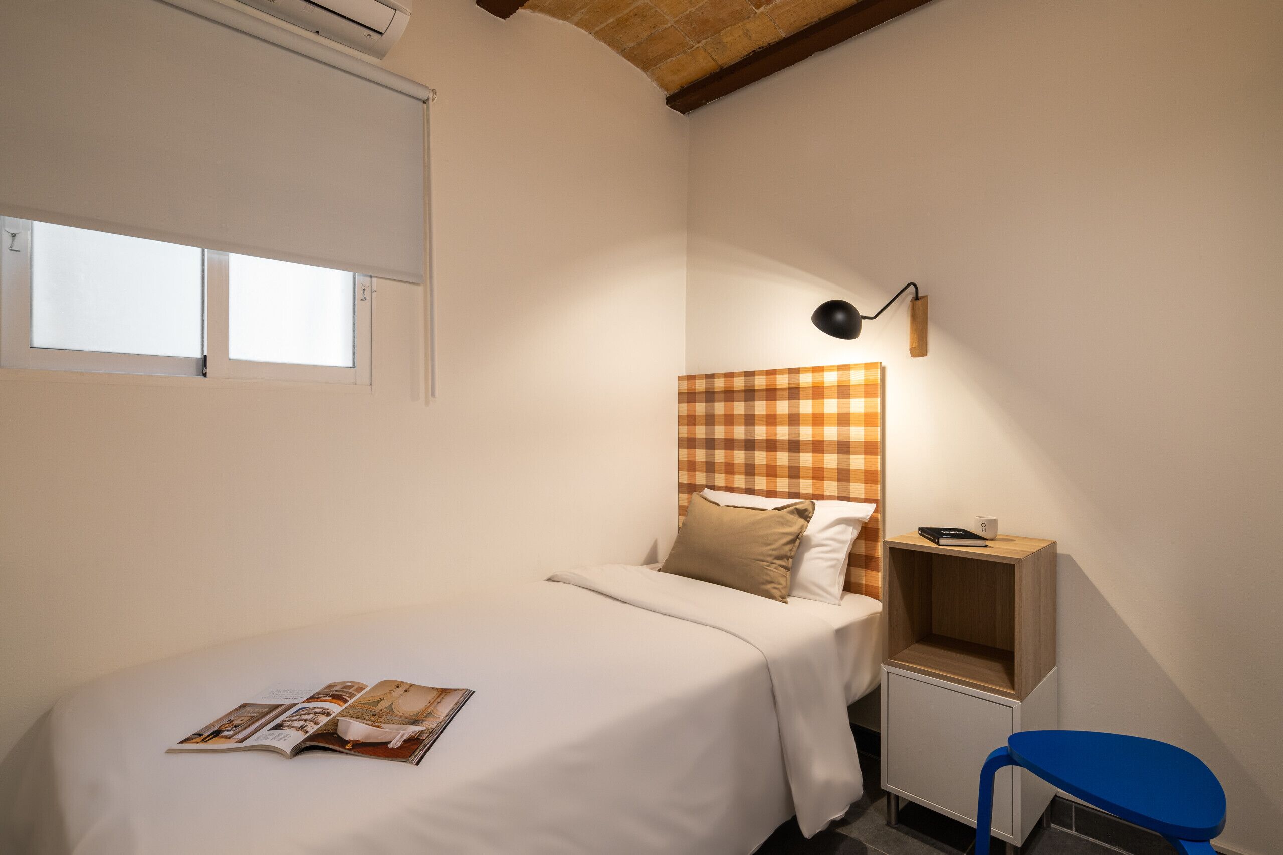 A bedroom with a bed, a nightstand, a chair, a checkered headboard, and a magazine on the bed.