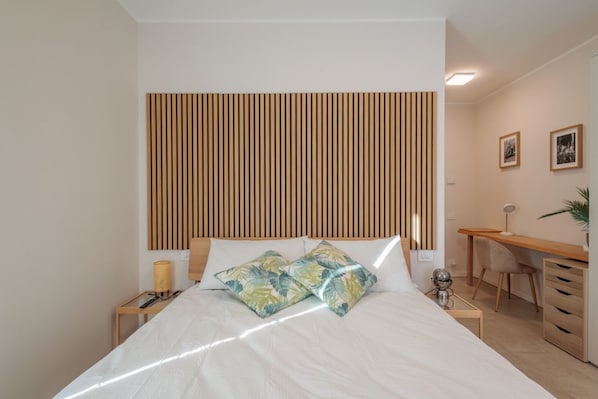 A bedroom with a bed, bedside table, lamp, and a desk with a chair.