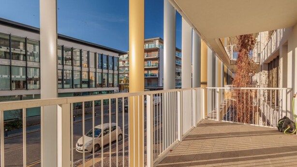 A balcony with a view of a street, a parked car, and a building with multiple floors and balconies.