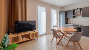 A modern living room with a flat-screen TV, a wooden entertainment unit, a dining table with chairs, and a kitchen area with cabinets and a stove.