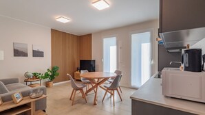 A modern living room with a sofa, TV, dining table, and chairs. There is a kitchen area with a microwave and a kettle.
