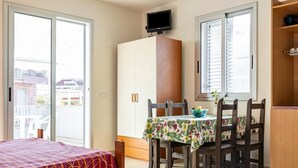 A room with a bed, a dining table with chairs, a TV on a cabinet, and a window with blinds. - GuestHost - Cozy 25 sqm studio apartment, perfect for 2 people, located on the first floor of a renovated building without a lift. The property is located in the center of Marina di Ragusa (20 km from Ragusa), one of the most renowned seaside areas in Si (Marina di Ragusa)