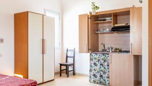 Une pièce de taille modeste comprenant un lit, une armoire en bois, une petite kitchenette avec un placard et une chaise.