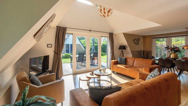 A spacious living room with a large sofa, a coffee table, and a TV. There is a sliding glass door leading to a patio with outdoor furniture. - Holiday Home Groote Keeten near Beaches (Groote Keeten)
