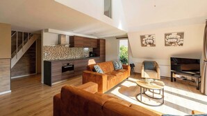 A modern living room with a sofa set, a coffee table, and a TV. There is a kitchen area with a countertop and cabinets. A staircase is visible in the background. - Holiday Home Groote Keeten near Beaches (Groote Keeten)