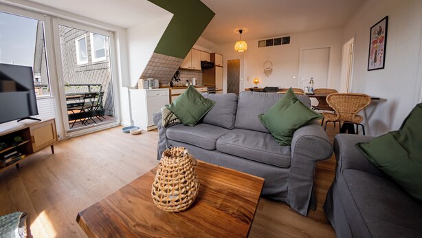 A modern living room with a grey sofa, wooden coffee table, and a wicker basket. A kitchen is visible in the background with a dining area. - "Küstenruhe" - your retreat at the Deichkieker guesthouse - pets allowed (Wangerland)