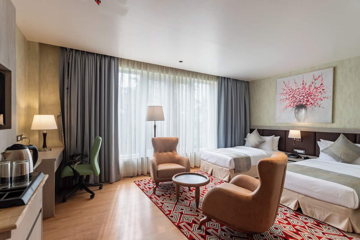 Quarto de hotel com uma cama, duas poltronas, uma escrivaninha com luminária, uma cafeteira e um quadro na parede.