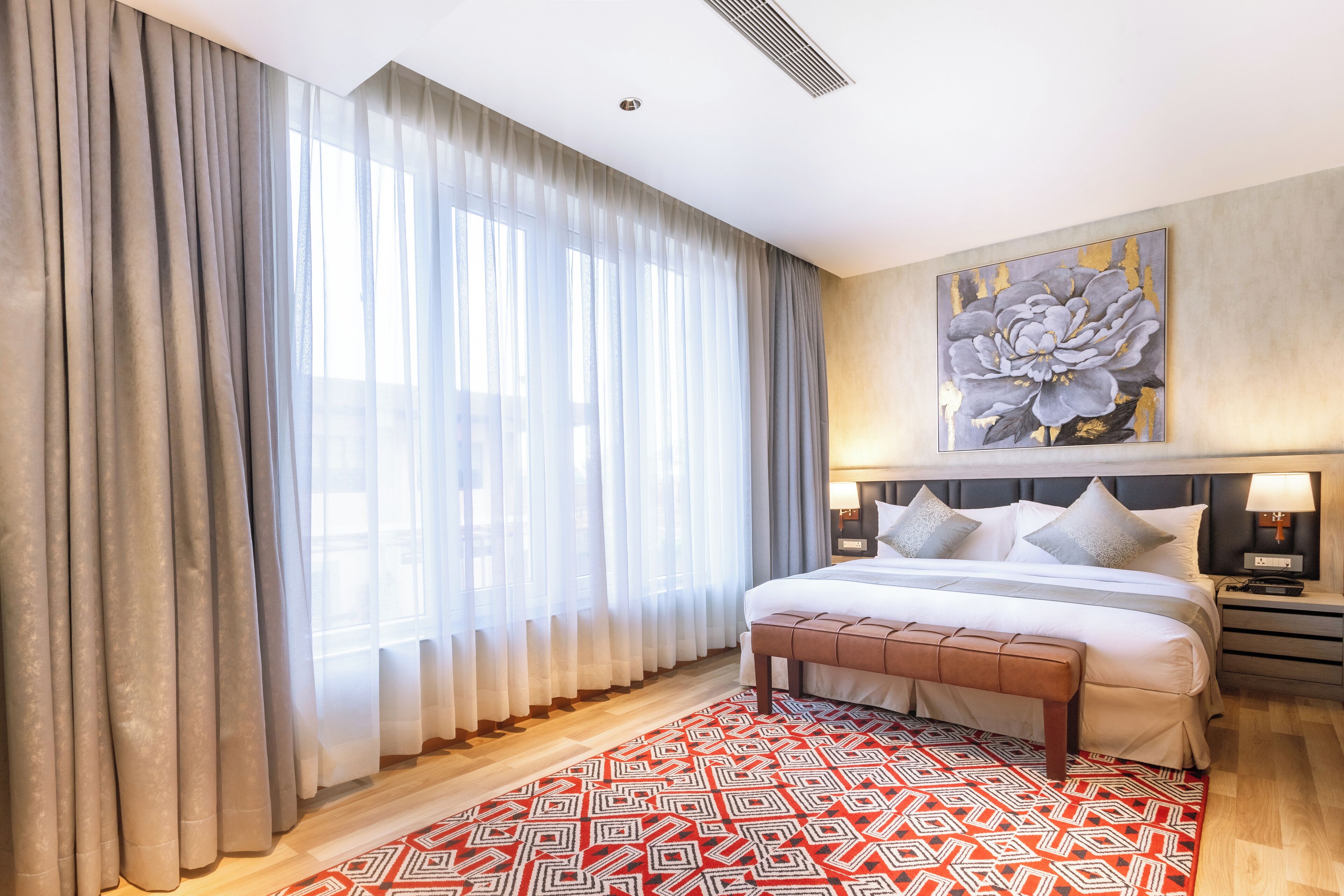 A modern hotel room with a large bed, a wooden floor, a red patterned rug, and a large floral artwork on the wall.