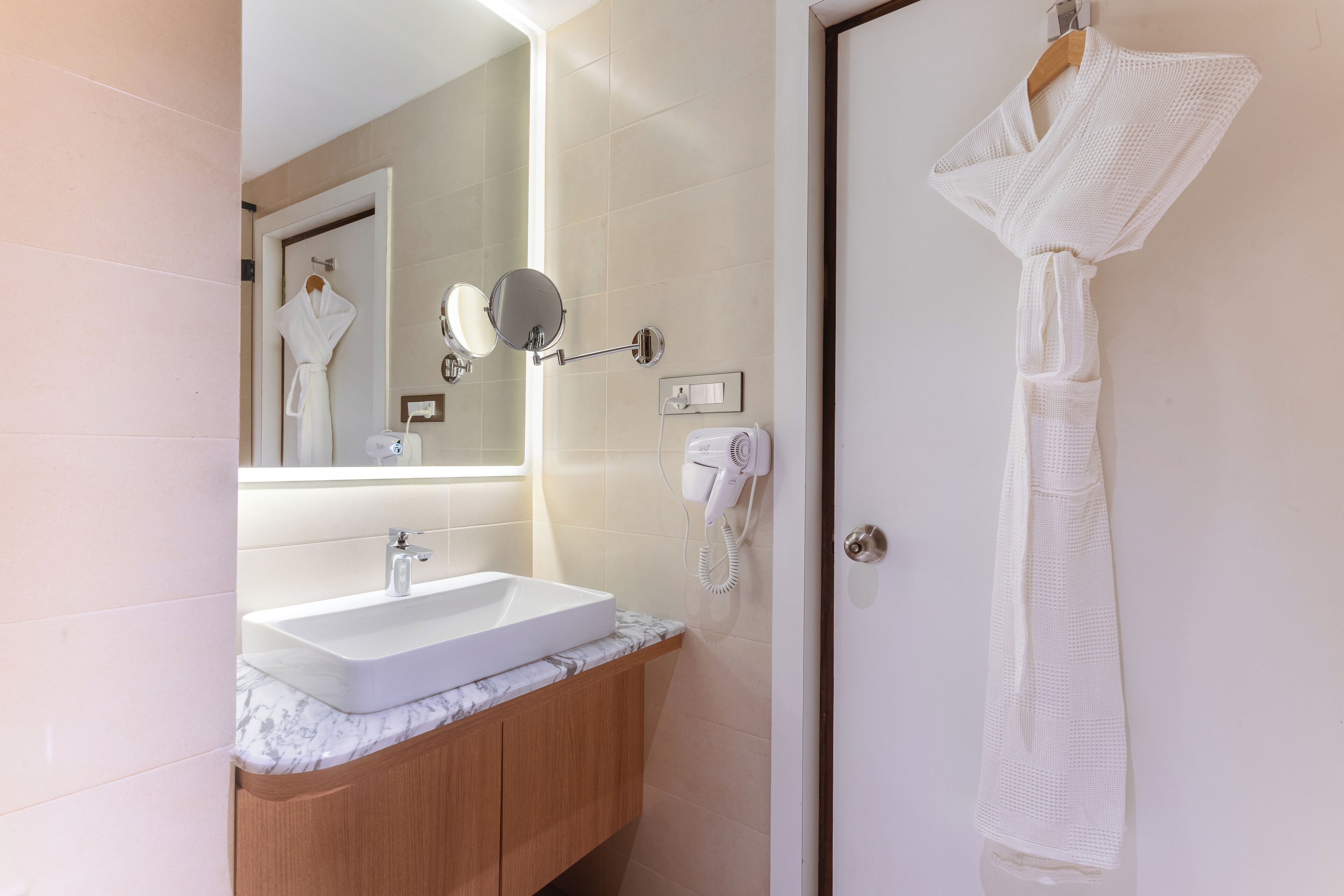 A modern bathroom with a marble sink, wooden cabinets, a mirror, and a white robe hanging on a door.