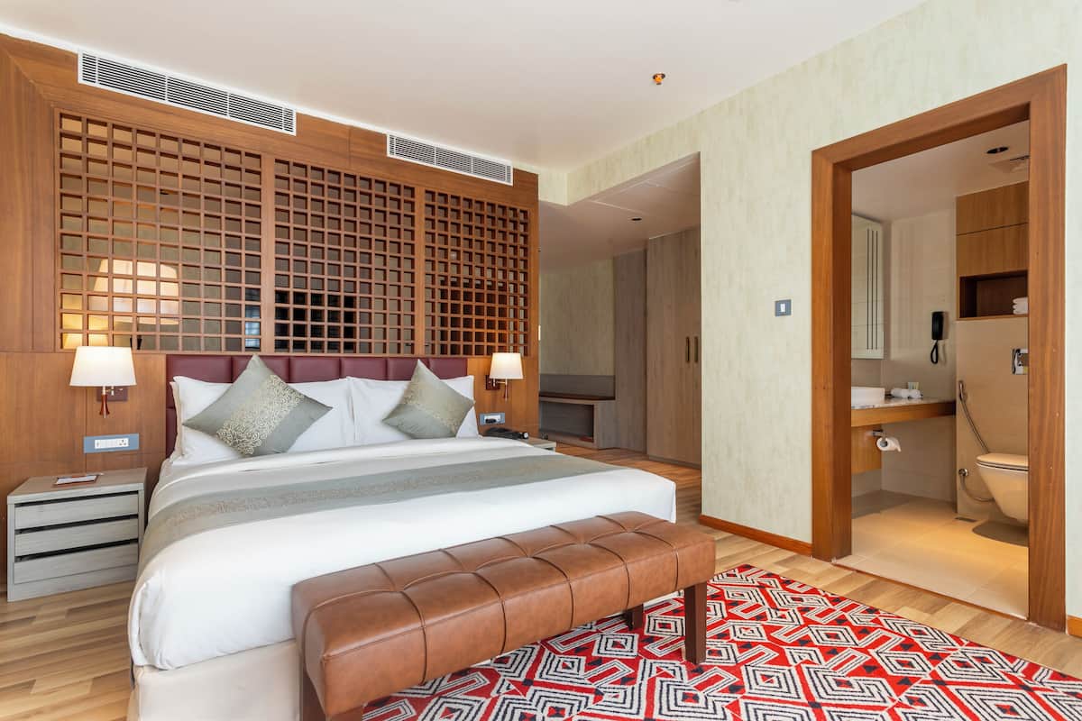 Quarto de hotel moderno com uma cama grande, cabeceira de madeira, um banco, um tapete estampado e um banheiro visível através de uma porta aberta.