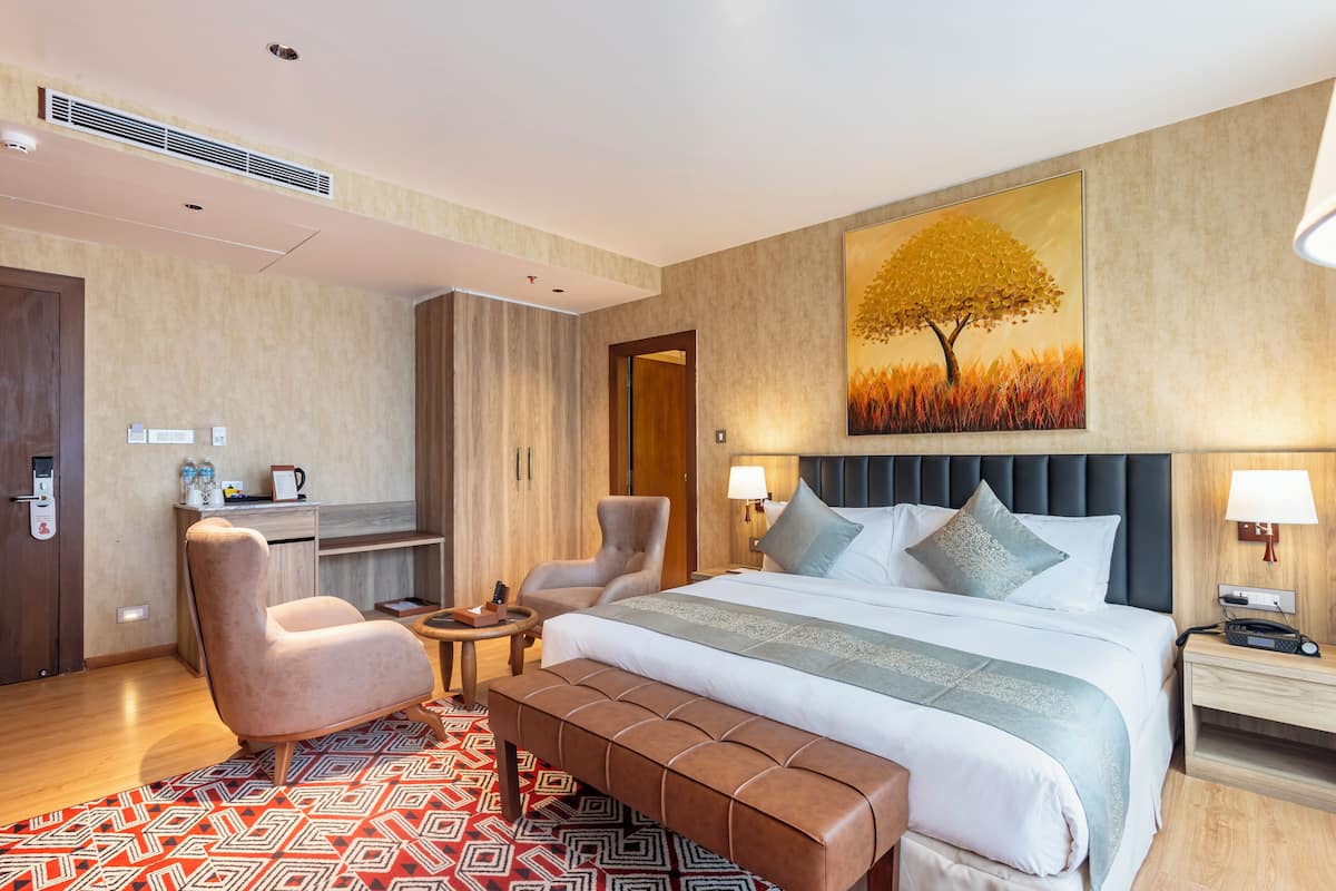 Quarto de hotel com uma cama grande, duas poltronas, uma mesinha, um banco, uma escrivaninha com cadeira e um quadro de uma árvore.