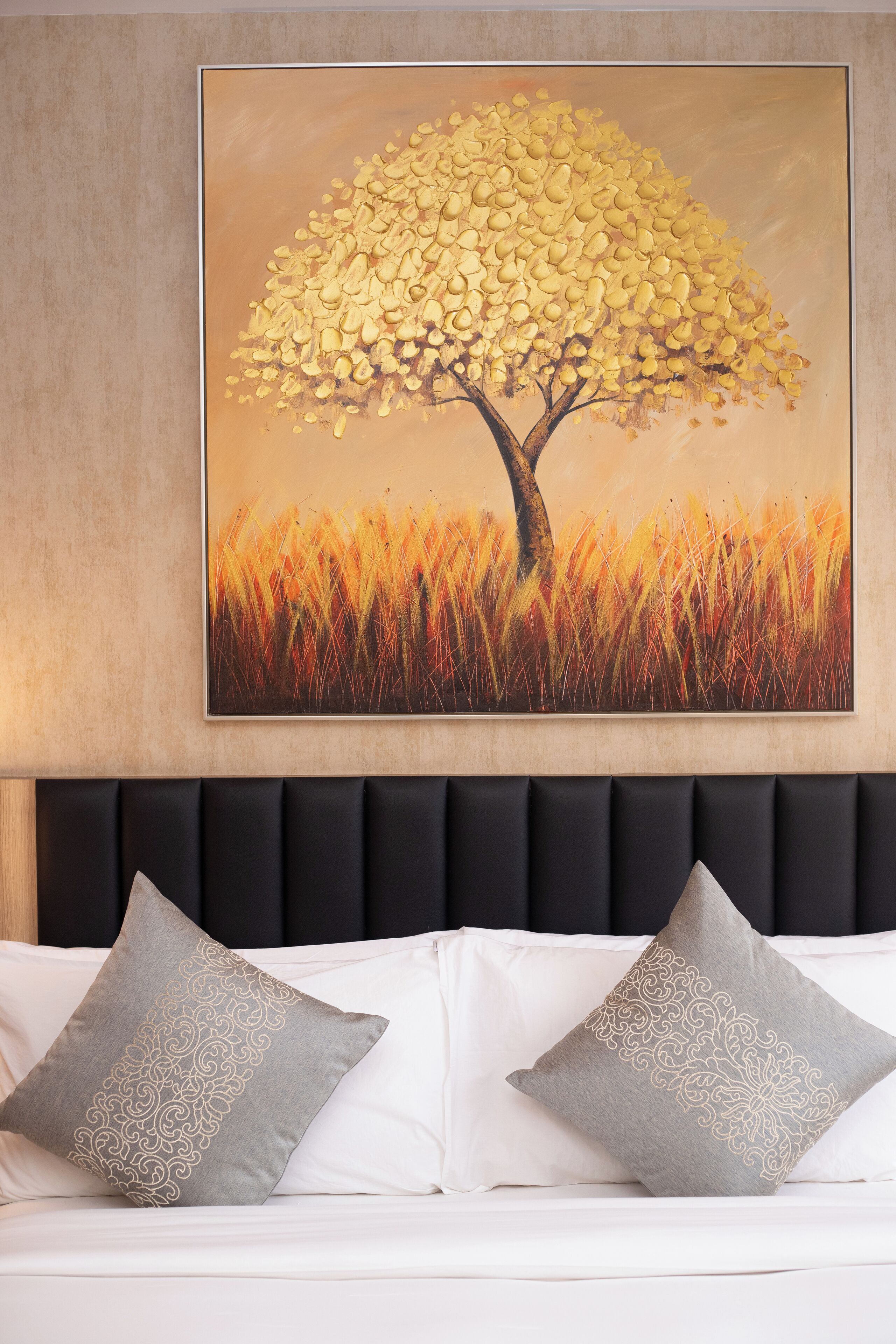 A room with a large painting of a tree on the wall, a black headboard, and two pillows with intricate patterns.