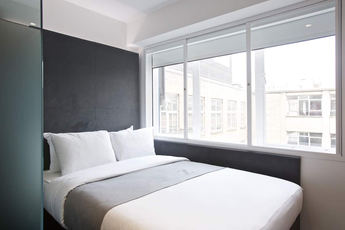 Una habitación de hotel moderna con una cama grande, una pared de acento oscura y vistas a edificios a través de un ventanal.