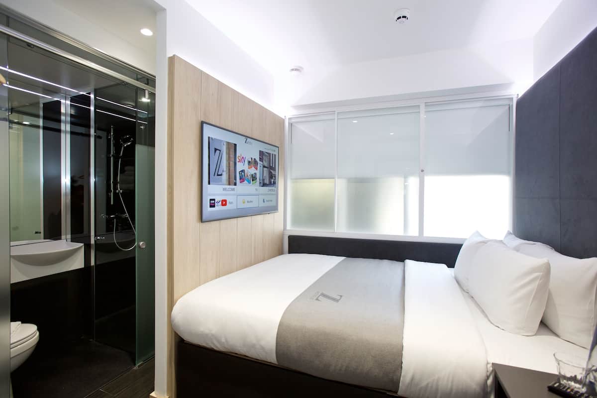 Una habitación de hotel moderna con una cama grande, un televisor montado, cabina de ducha con mampara de vidrio y un ventanal amplio.