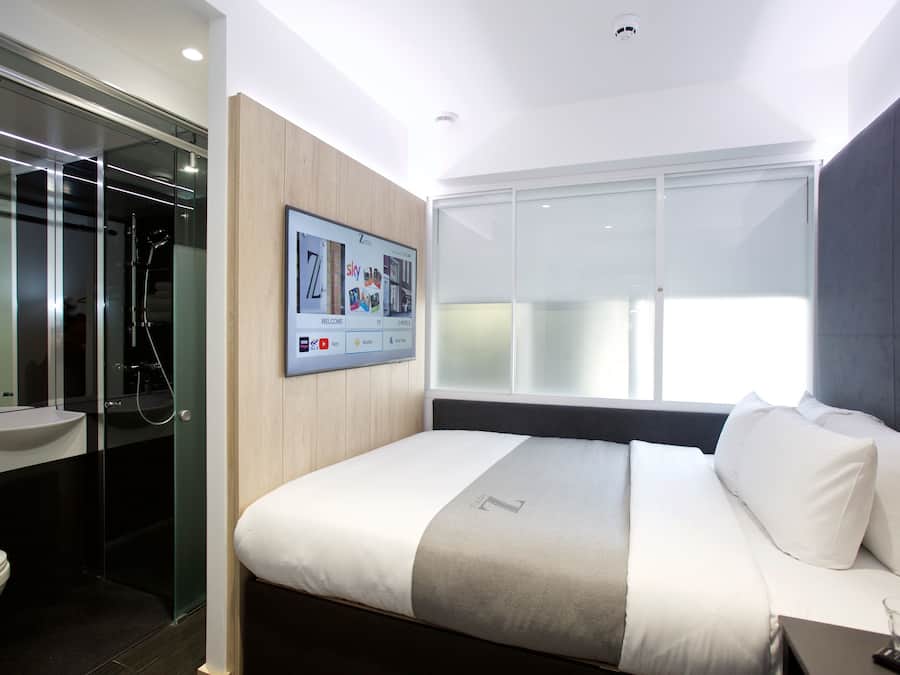 Ein modernes Hotelzimmer mit einem großen Bett, einem wandmontierten Fernseher, einer Duschkabine aus Glas und einem großen Fenster.