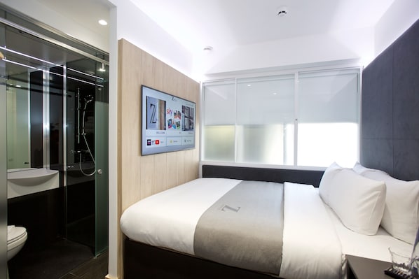 Una habitación de hotel moderna con una cama grande, un televisor montado, cabina de ducha con mampara de vidrio y un ventanal amplio.