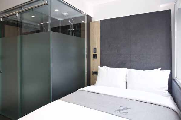 A modern hotel room with a glass partition, a grey headboard, and a bed with white linens.