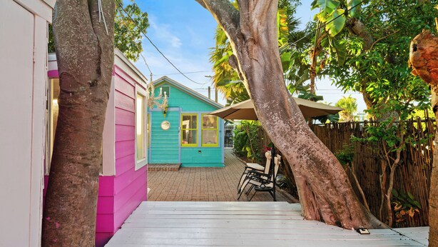 Un étroit sentier en bois mène à une maison turquoise dotée d’une véranda, entourée d’arbres et d’une clôture en bambou.