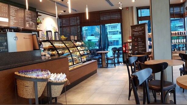 L’intérieur d’un café présente un comptoir où sont exposées des pâtisseries, un tableau affichant le menu, ainsi qu’un espace avec des tables et des chaises.