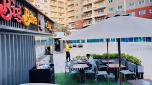 Un café en plein air avec des tables et des chaises, une personne debout près de l’entrée et un bâtiment avec une enseigne en arrière-plan.