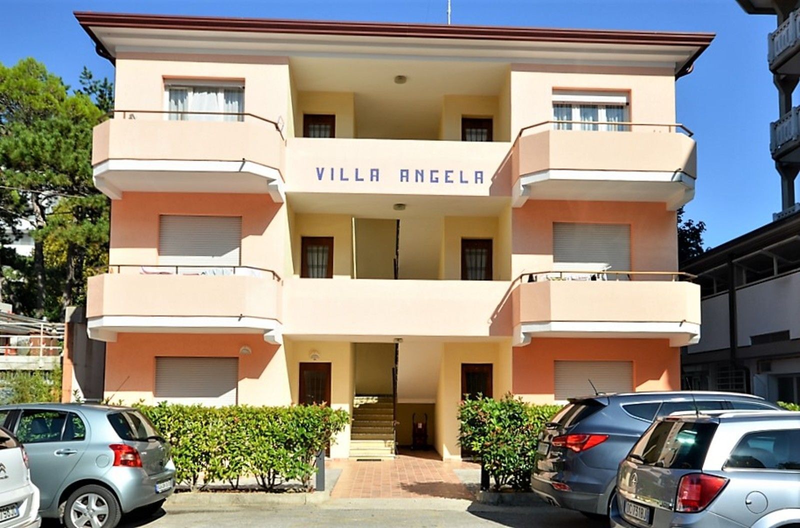 Ein mehrstöckiges Gebäude namens "Villa Angela" mit einem zentralen Eingang, Balkonen und parkenden Autos davor.