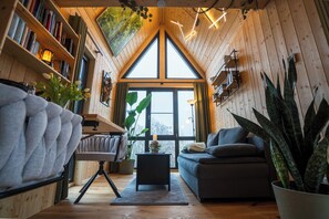 Un interior acogedor de madera con un ventanal grande, un sofá, una estantería y varias plantas.