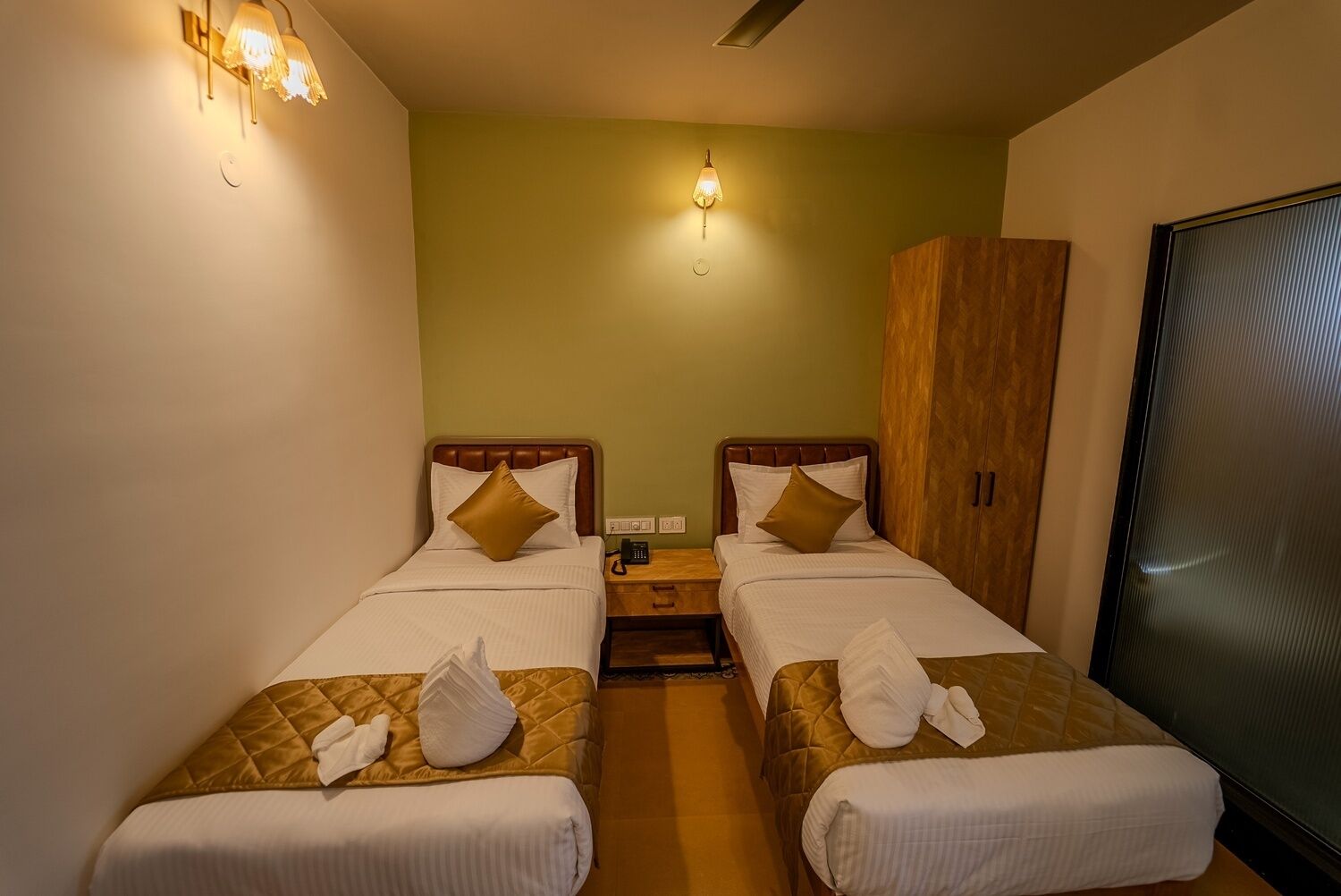 A hotel room with two beds, a wardrobe, a bedside table with a phone, and a wall-mounted light fixture.