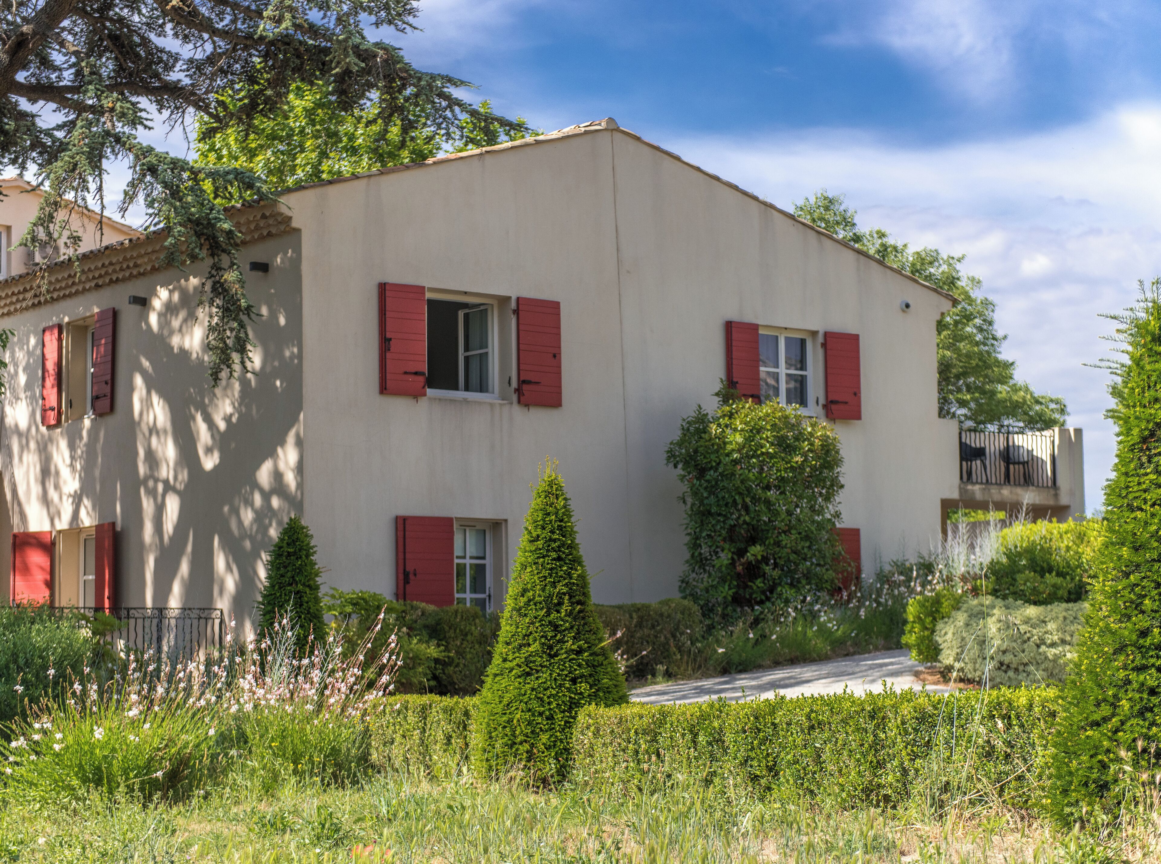 A two-story building with red shutters, surrounded by greenery and a clear sky.