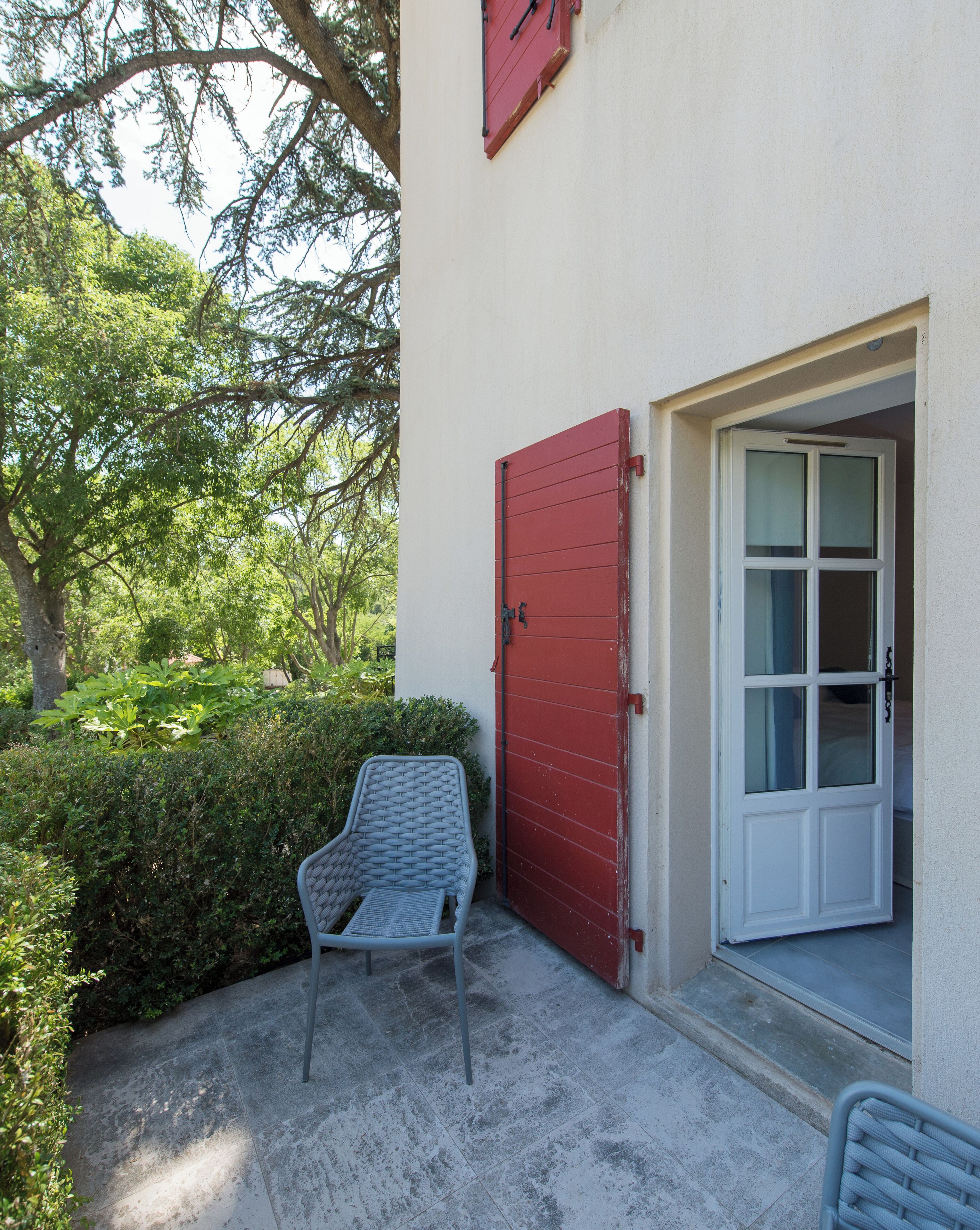 Un edificio con puerta roja y ventana blanca, una silla afuera y follaje al fondo.