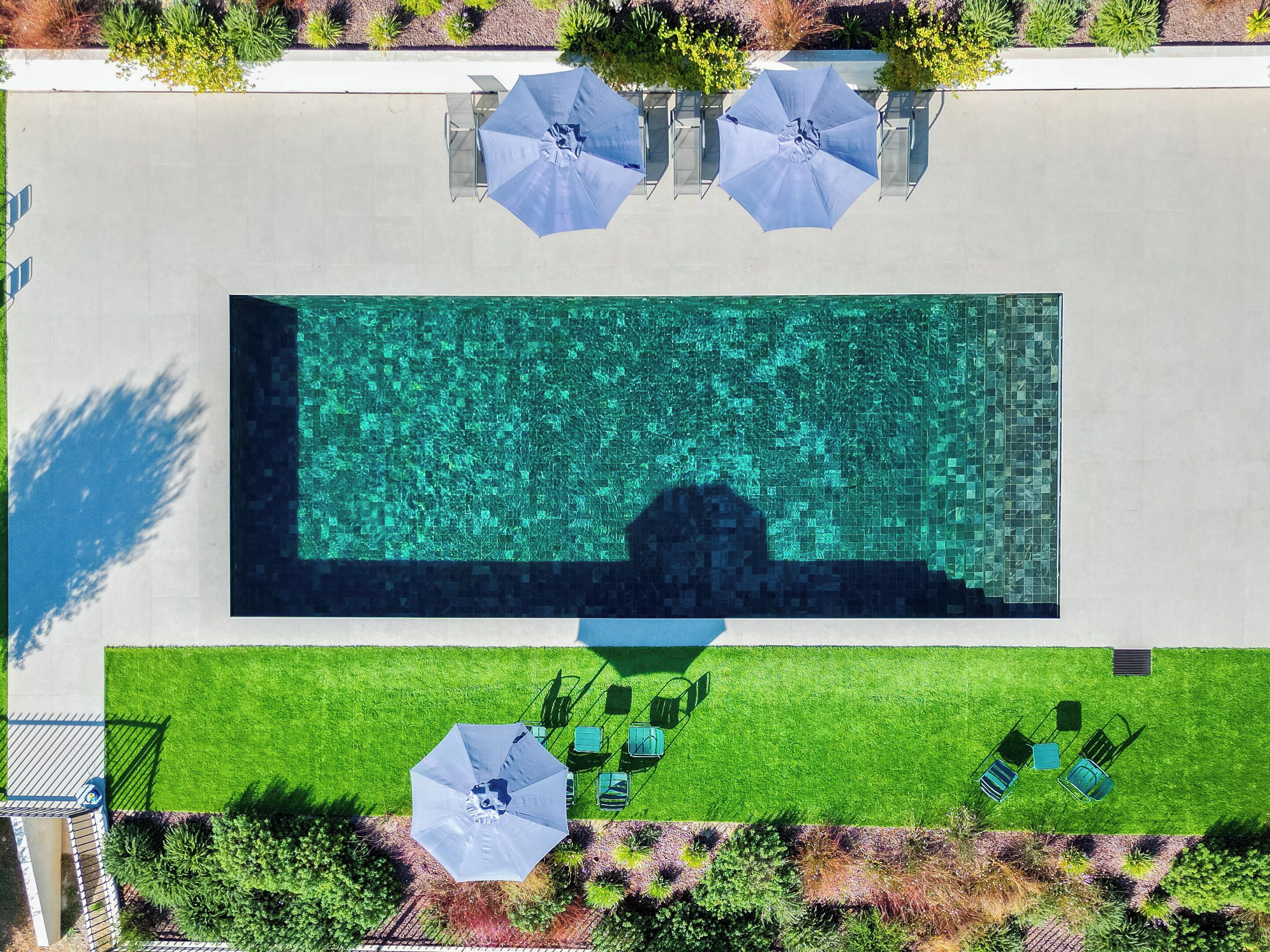 Vista aérea de una piscina rectangular con un deck o solárium empedrado alrededor. Hay tres sombrillas y varias sillas de descanso dispuestas junto a la piscina.