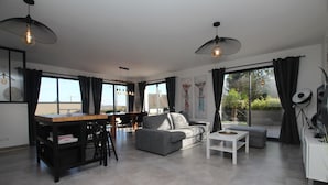 Una sala de estar moderna con sofá, mesa de centro y comedor con sillas. Hay ventanas grandes con cortinas, obras de arte en las paredes y un ventilador de techo.