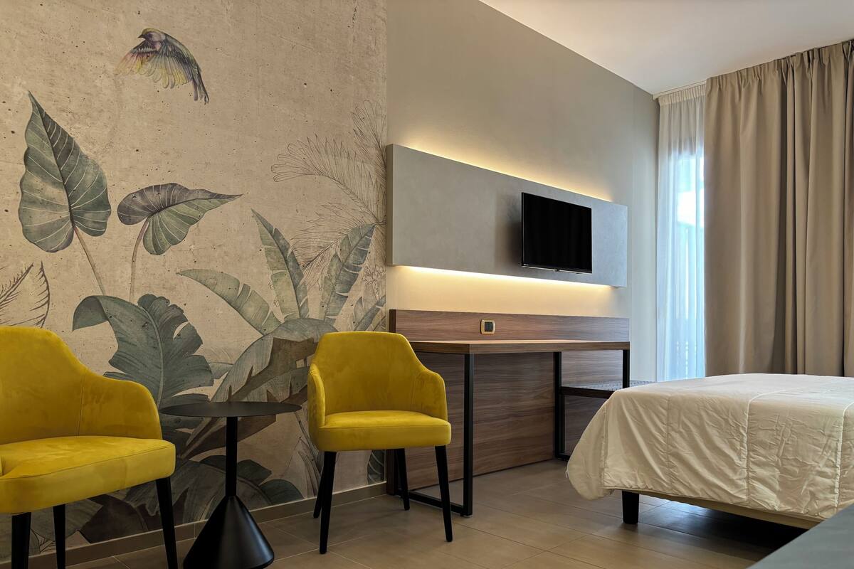 Une chambre d’hôtel avec un lit, deux fauteuils jaunes, une petite table, un téléviseur à écran plat et une grande fenêtre avec des rideaux.