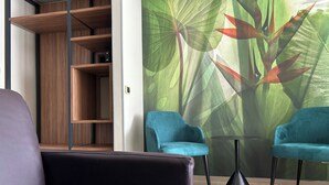 A modern room with a wooden shelving unit, two teal armchairs, a small round table, and a large wall mural of tropical leaves. - Casa Vacanze Fardella Centrale (Trapani)