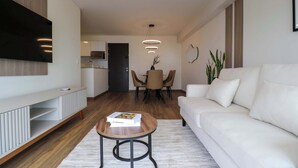 A modern living room with a white sofa, a wooden coffee table with a book, a TV mounted on the wall, a dining area with a table and chairs, and a kitchen in the background. - w* | Pleasant 3BR w/ Balcony in San Isidro (Lima)