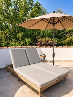 Een ligbed met een gevlochten frame en lichtgrijze kussens, onder een grote beige parasol op een betegeld terras.