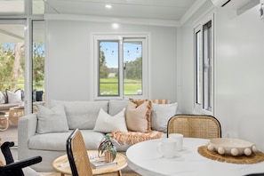 Relax in this serene living room with a light grey sofa, wicker chair, and round coffee table, surrounded by lush garden views.