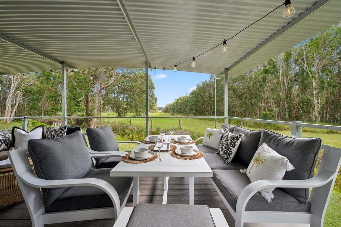 Un espace extérieur aménagé avec une table pour deux, entouré de mobilier blanc agrémenté de coussins gris, sous un toit abrité illuminé de guirlandes lumineuses.