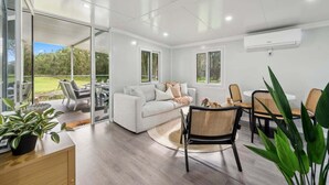 A modern living room with a sofa, chairs, a dining table, and a view of the outdoors. - Leos Beach Hideaway, Stylish Tiny Home (Coffs Harbour)