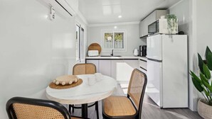 A compact kitchen with white appliances, a dining table with chairs, and a window. - Leos Beach Hideaway, Stylish Tiny Home (Coffs Harbour)
