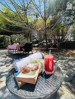 Una mesa con un libro, gafas de sol y una bebida, ubicada al aire libre con árboles y un cielo despejado de fondo.