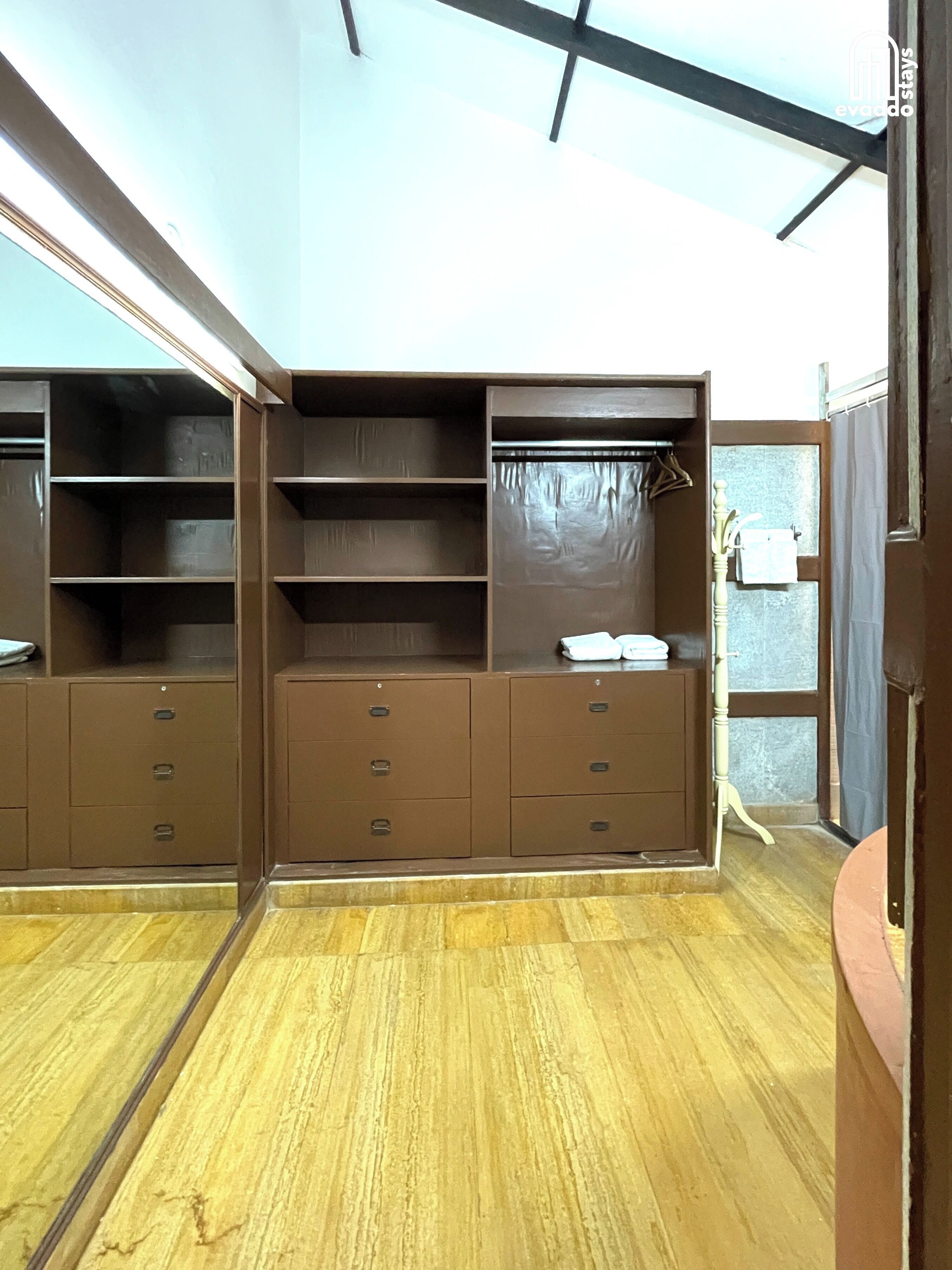 A room with a wooden floor, a brown wardrobe with multiple drawers and shelves, and a door with a towel hanging on it.