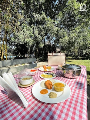 A picnic setup with a checkered tablecloth, various dishes, and a portable stove on a grassy area surrounded by trees. - The Hideaway Hosur by evaddo stays (Hosur)