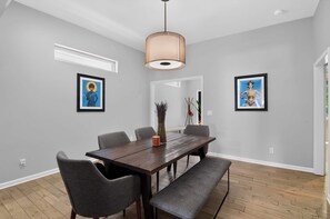A dining area with a wooden table, grey upholstered chairs, a vase with dried plants, and framed artwork on the walls.