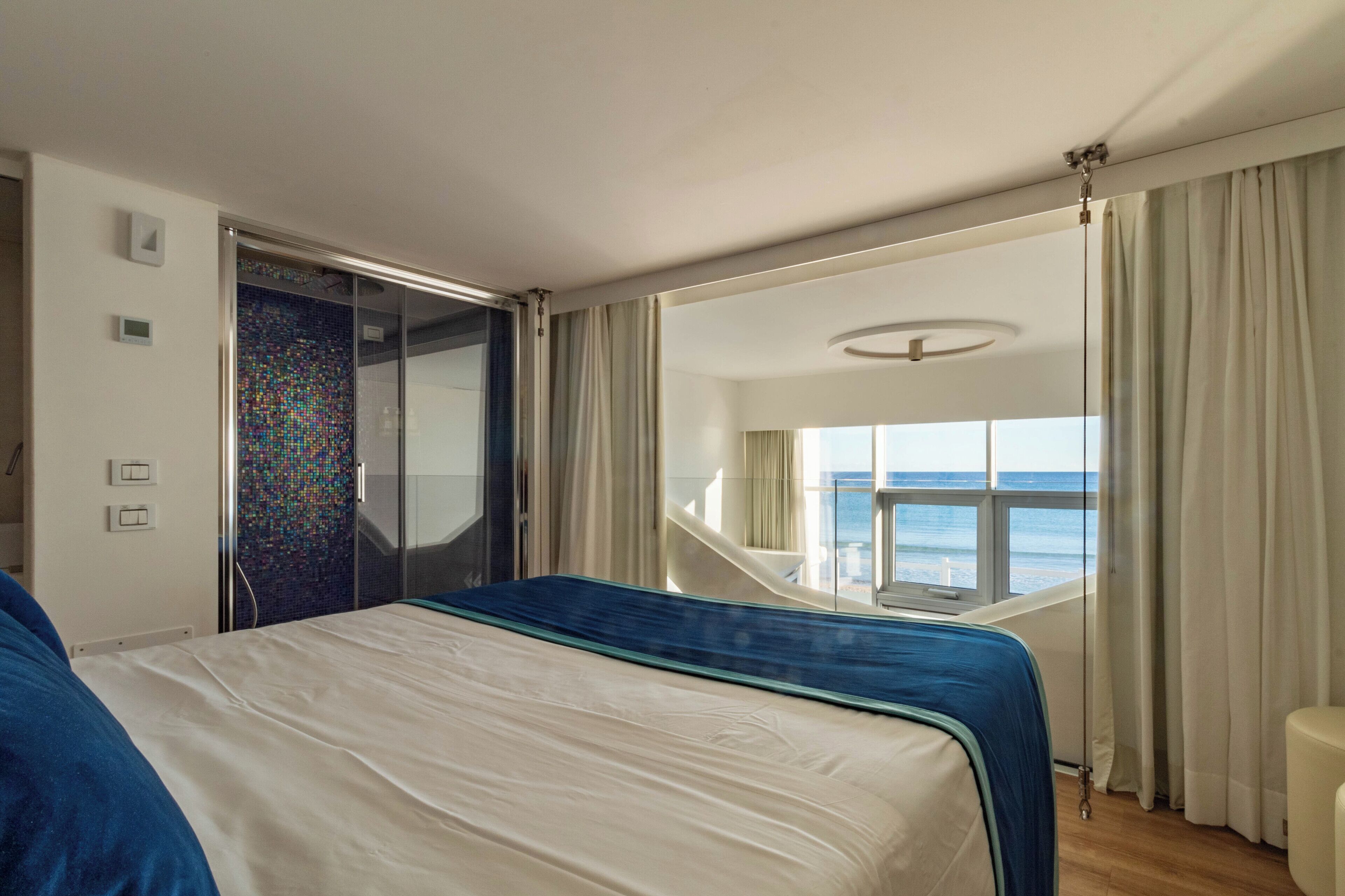 Quarto de hotel com duas camas, uma varanda e vista para o mar, e uma janela grande com cortinas.