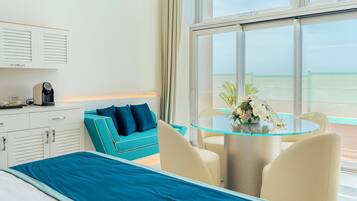 Une chambre d’hôtel avec un lit, un canapé de couleur turquoise, une table en verre avec des chaises, une machine à café et une vue sur la mer par de grandes fenêtres.