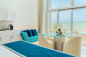 Habitación de hotel con cama, sofá color turquesa, mesa de vidrio con sillas, cafetera y vista al mar a través de amplias ventanas.
