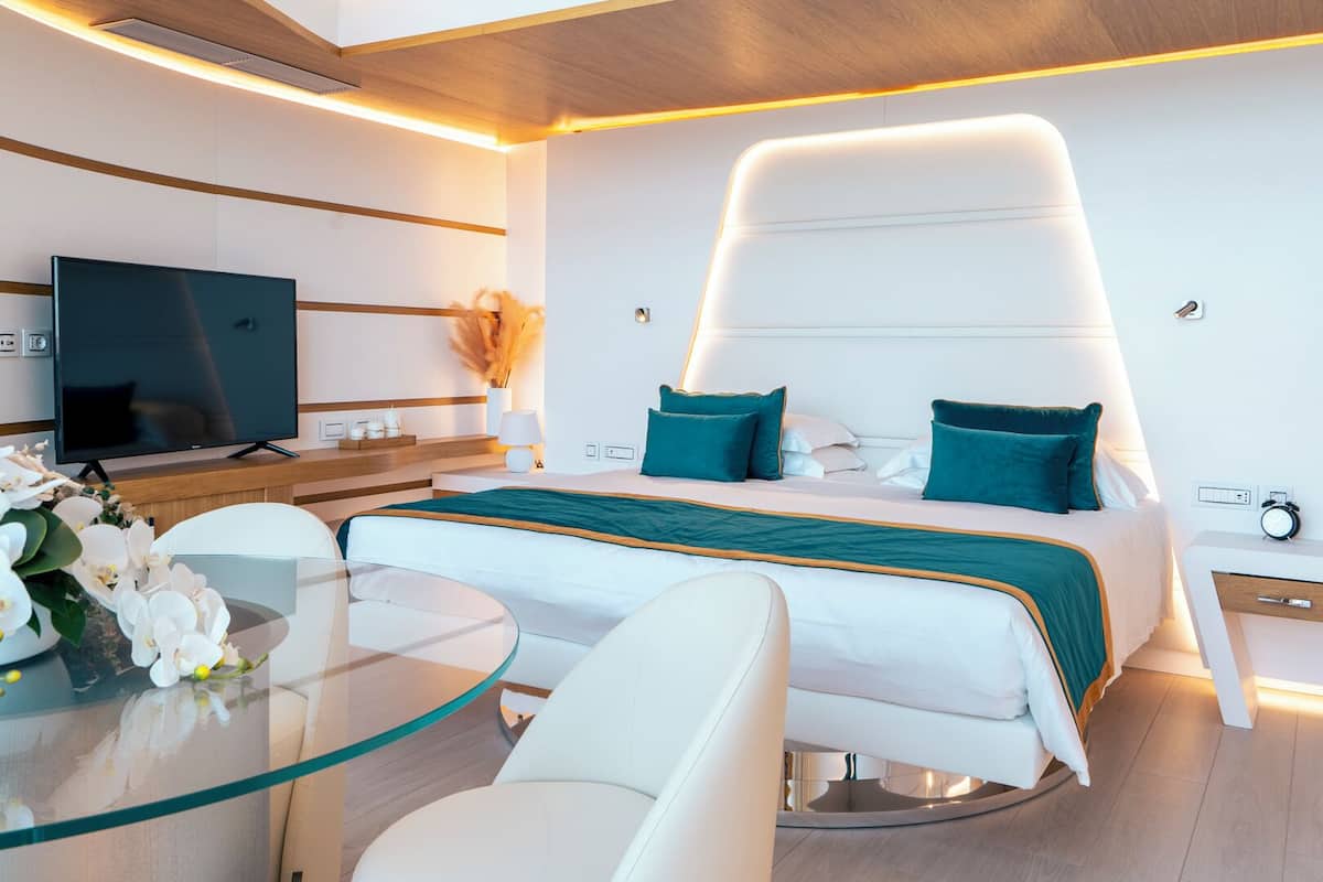 A modern hotel room with a bed, a glass table, white chairs, a TV, and a bedside table with a clock.