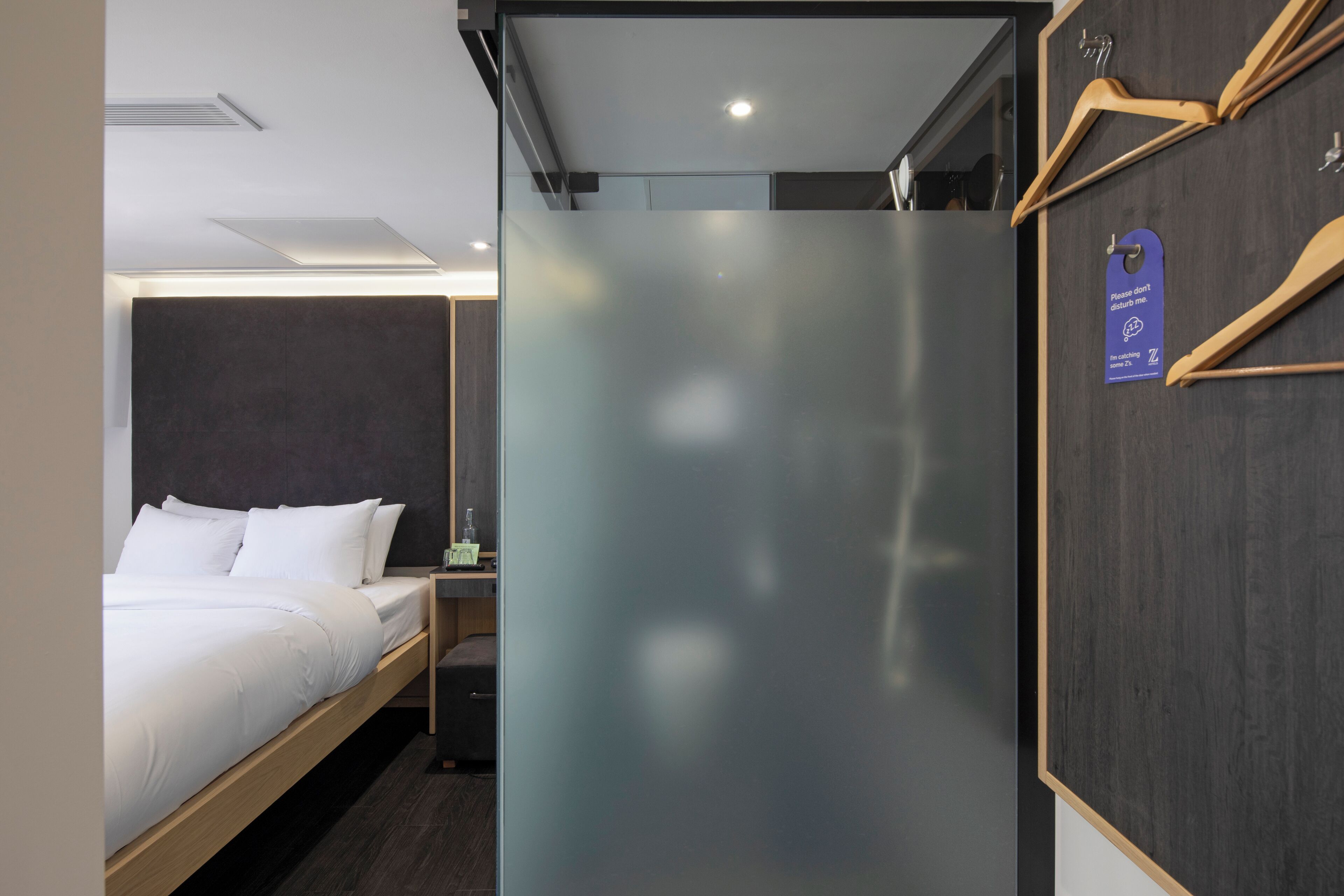 Ein Hotelzimmer mit Bett, Nachttisch, Spiegel und einem Kleiderhaken aus Holz mit einem Etikett.